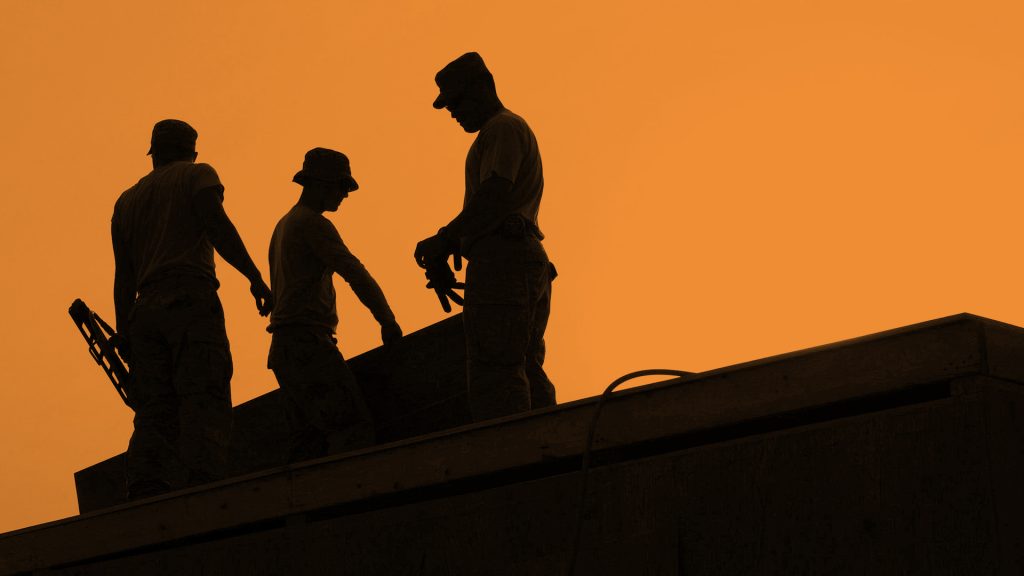 Construction workers on a roof, they are silhouetted by the sun behind them. The entire photo has an orange tint overlay.