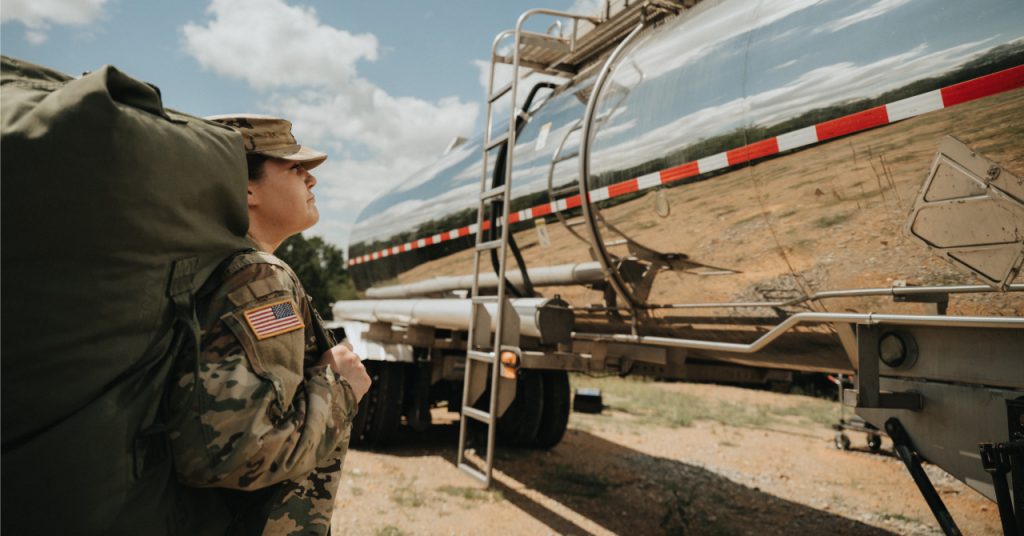 A woman in military gear looking at her reflection on the side of a tank truck 