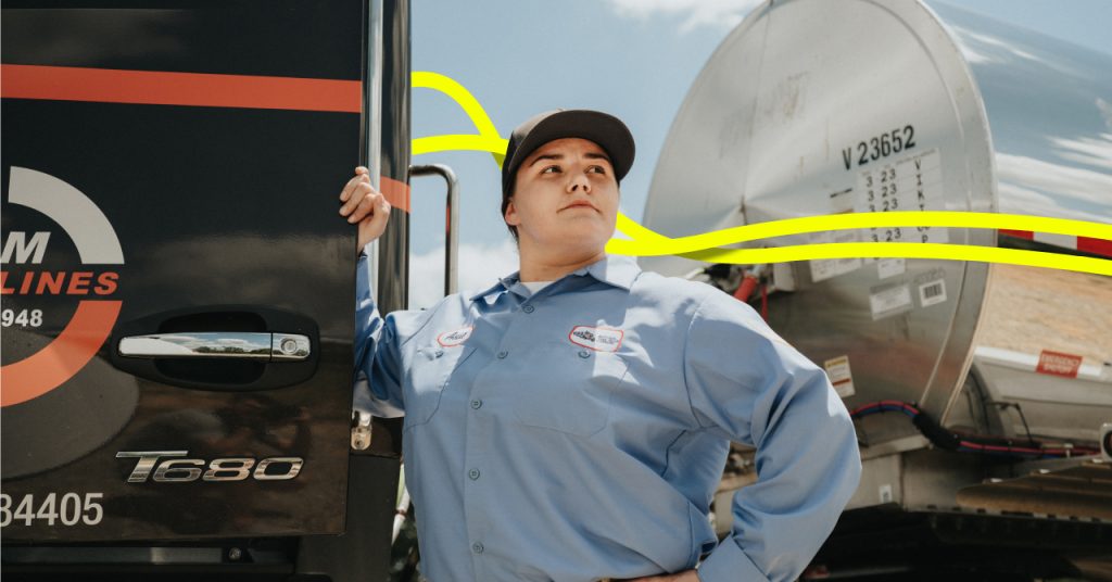 A woman in a trucking uniform looking into the distance while leaning on the back of a tank truck